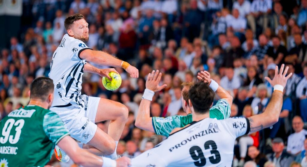 Ein Handballspieler in einem weißen Trikot springt in der Luft, um den Ball zu schießen, während zwei Verteidiger in grünen Trikots ihre Arme heben, um ihn zu blockieren. Eine große Menschenmenge beobachtet das intensive Spiel im Hintergrund.
