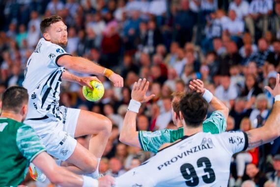 Ein Handballspieler in einem weißen Trikot springt in der Luft, um den Ball zu schießen, während zwei Verteidiger in grünen Trikots ihre Arme heben, um ihn zu blockieren. Eine große Menschenmenge beobachtet das intensive Spiel im Hintergrund.
