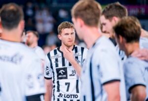 Eine Gruppe männlicher Handballspieler in weißen und schwarzen Trikots versammelt sich in einem Huddle. Ein Spieler, der die Nummer 7 trägt, steht in der Mitte, berührt sein Kinn und schaut nachdenklich, während er seinen Mitspielern zuhört.