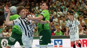 Zwei Männer kämpfen während eines Handballspiels um den Ball, der eine in einem weiß-schwarz gestreiften Trikot, der andere in einem grünen Trikot, während die Zuschauer im Hintergrund zusehen.