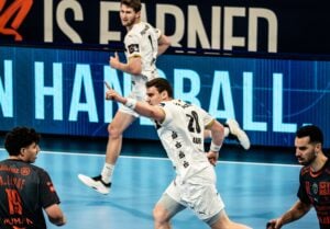 Ein Handballspieler in einem weißen Trikot rennt und zeigt nach vorne, um auf einem Hallenplatz zu jubeln, während andere Spieler in weißen und dunklen Trikots im Hintergrund zu sehen sind. Auf einer blauen LED-Anzeige steht HANDBALL.
