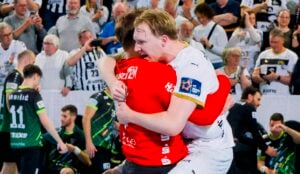 Zwei männliche Handballspieler in rot-weißen Trikots umarmen sich freudig auf dem Spielfeld, während im Hintergrund eine jubelnde Menge und andere Spieler zu sehen sind.