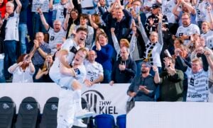 Zwei Sportler in weißen Trikots feiern ein Tor; ein Spieler hebt den anderen unter Jubel hoch. Eine Menge begeisterter Fans in passenden Trikots und Schals jubelt ihnen in einer Sportarena begeistert zu.
