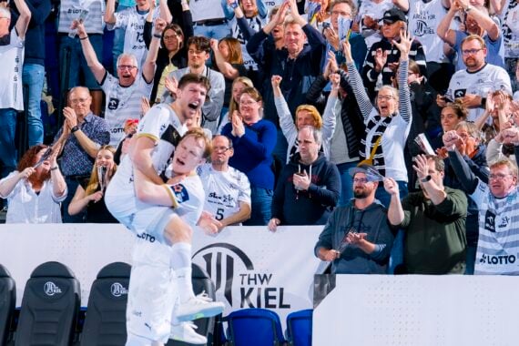 Zwei Sportler in weißen Trikots feiern ein Tor; ein Spieler hebt den anderen unter Jubel hoch. Eine Menge begeisterter Fans in passenden Trikots und Schals jubelt ihnen in einer Sportarena begeistert zu.