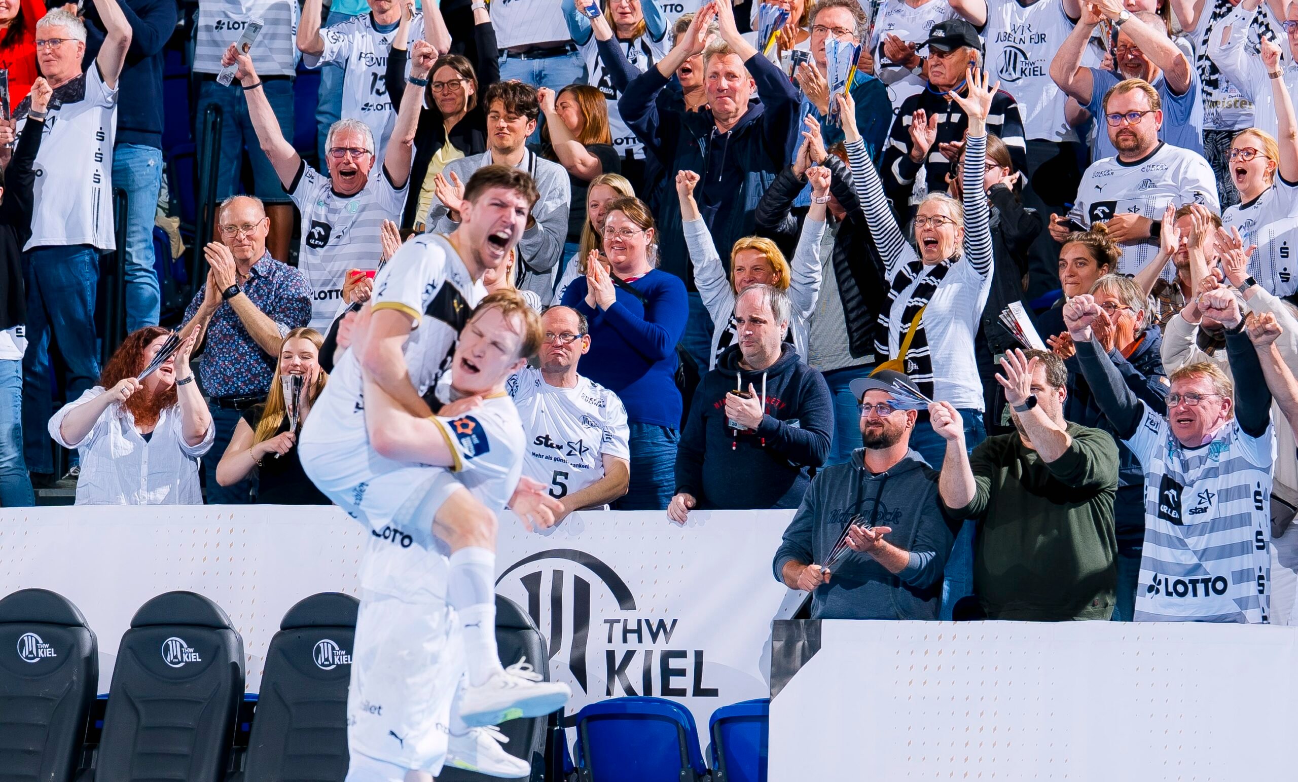 Zwei Sportler in weißen Trikots feiern ein Tor; ein Spieler hebt den anderen unter Jubel hoch. Eine Menge begeisterter Fans in passenden Trikots und Schals jubelt ihnen in einer Sportarena begeistert zu.
