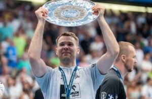 Ein männlicher Sportler in einem weißen Trikot hält lächelnd eine große silberne Trophäenplatte über seinen Kopf, während er eine Medaille um den Hals trägt und die Menge im Hintergrund jubelt.