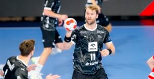 Ein männlicher Handballspieler in einem schwarz-weißen Trikot hält einen Handball und bereitet sich darauf vor, ihn während eines Spiels zu werfen. Andere Spieler sind im Hintergrund auf einem blauen Hallenplatz zu sehen.