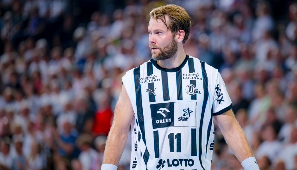 Ein männlicher Handballspieler in einem weiß-schwarz gestreiften Trikot mit der Nummer 11 steht auf dem Spielfeld und schaut nach links. Eine unscharfe Menschenmenge füllt den Hintergrund.