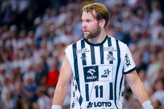 Ein männlicher Handballspieler in einem weiß-schwarz gestreiften Trikot mit der Nummer 11 steht auf dem Spielfeld und schaut nach links. Eine unscharfe Menschenmenge füllt den Hintergrund.