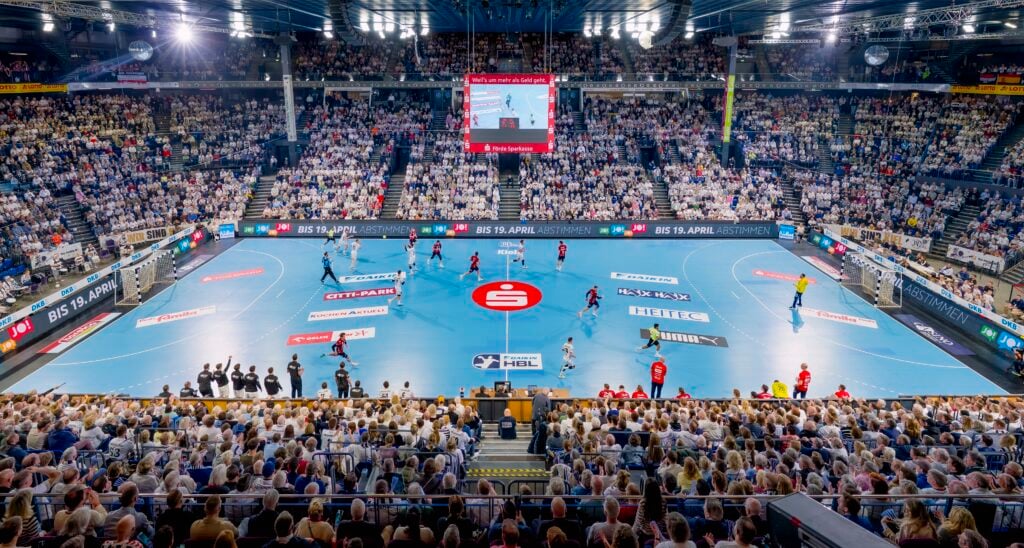 Weite Sicht auf eine überfüllte Halle während eines Handballspiels, mit Spielern auf einem blauen Spielfeld, einer Anzeigetafel darüber und Zuschauern auf den Tribünen. Helle Lichter erhellen die Szene.
