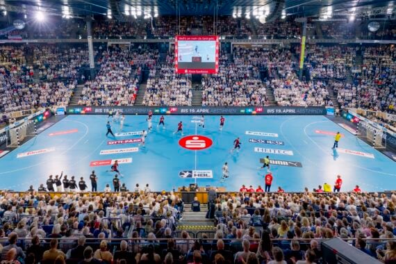 Weite Sicht auf eine überfüllte Halle während eines Handballspiels, mit Spielern auf einem blauen Spielfeld, einer Anzeigetafel darüber und Zuschauern auf den Tribünen. Helle Lichter erhellen die Szene.