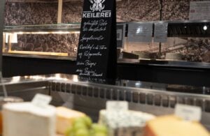 Im Vordergrund eine Vitrine mit verschiedenen Käsesorten, hinter der Theke eine Speisekarte mit handgeschriebenen Einträgen in deutscher Sprache und an der Rückwand ein Wandfoto mit einer jubelnden Menge.