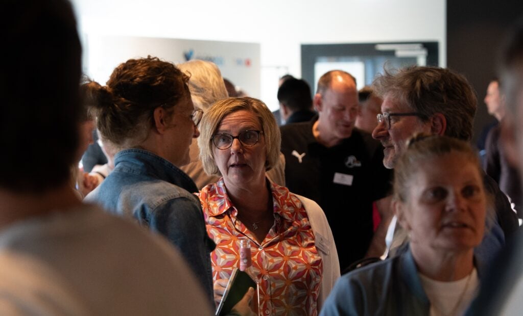Eine Gruppe von Menschen versammelt sich in einem Innenraum und unterhält sich in kleinen Gruppen. Eine Frau mit Brille und bunter Bluse steht im Mittelpunkt und unterhält sich mit einer anderen Frau in einer Jeansjacke. Es scheint sich um eine gesellschaftliche oder berufliche Veranstaltung zu handeln.