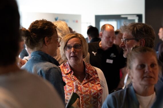 Eine Gruppe von Menschen versammelt sich in einem Innenraum und unterhält sich in kleinen Gruppen. Eine Frau mit Brille und bunter Bluse steht im Mittelpunkt und unterhält sich mit einer anderen Frau in einer Jeansjacke. Es scheint sich um eine gesellschaftliche oder berufliche Veranstaltung zu handeln.