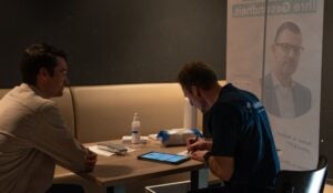 Zwei Männer sitzen sich an einem Tisch gegenüber; einer benutzt ein Tablet, während der andere zuhört. Auf dem Tisch befinden sich eine Flasche Desinfektionsmittel, ein Gerät und Zubehör. Im Hintergrund ist ein Transparent mit dem Porträt eines Mannes zu sehen.