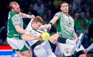 Zwei Handballspieler in grünen Trikots verteidigen sich aggressiv gegen einen Spieler in einem weißen Trikot und packen ihn am Hemd, während er versucht, den gelben Ball zu schießen. Die Aktion wirkt intensiv und körperlich auf einem Hallenplatz.