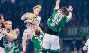 Ein Handballspieler in einem weißen Trikot springt mit dem Ball in der Luft, um ihn zu werfen, während drei Verteidiger in Grün versuchen, ihn während eines intensiven Spiels in einer Halle zu blockieren.