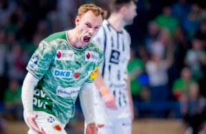 Ein männlicher Handballspieler in einem grünen Trikot schreit während eines Spiels energisch, während ein anderer Spieler in einem weiß-schwarzen Trikot im Hintergrund verschwommen zu sehen ist und eine jubelnde Menge hinter ihnen steht.