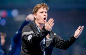 Ein Mann, der eine schwarze Sportjacke und ein Schlüsselband trägt, gestikuliert mit beiden Händen und scheint zu schreien oder Anweisungen zu geben, möglicherweise während einer Sportveranstaltung. Der Hintergrund ist unscharf, andere Personen sind zu sehen.