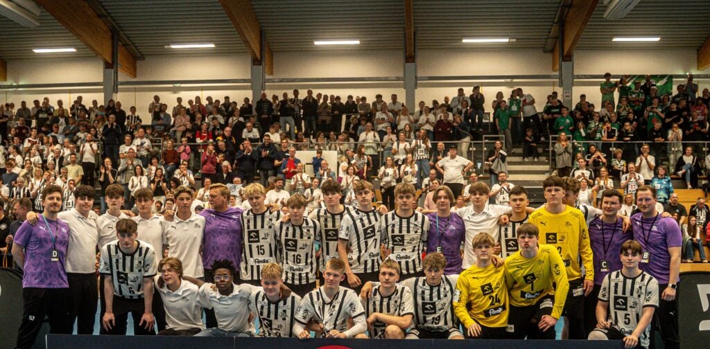 Eine große Gruppe junger männlicher Athleten in schwarz-weiß gestreiften Uniformen posiert für ein Mannschaftsfoto in einer Halle, umgeben von Trainern und Mitarbeitern und einer jubelnden Menge auf der Tribüne dahinter.