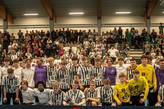 Eine große Gruppe junger männlicher Athleten in schwarz-weiß gestreiften Uniformen posiert für ein Mannschaftsfoto in einer Halle, umgeben von Trainern und Mitarbeitern und einer jubelnden Menge auf der Tribüne dahinter.
