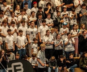 Eine große Gruppe von Fans, meist junge Leute, steht in einem Stadion dicht beieinander, viele tragen passende weiße und schwarze Sporttrikots, verfolgen aufmerksam ein Ereignis und klatschen oder jubeln.