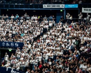 Eine große Menge von Fans, viele in weißen Hemden, füllen die Sitze einer Sporthalle. Auf Bannern steht WIR SIND KIEL. Einige Menschen stehen, während die meisten sitzen und das Geschehen aufmerksam verfolgen.
