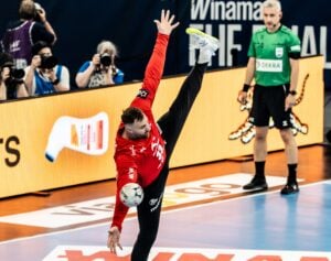 Ein Handballtorhüter in einem roten Trikot führt einen Fallrückzieher aus, indem er sein linkes Bein nach oben streckt, während er einen Ball abwehrt. Im Hintergrund sind Fotografen und ein Schiedsrichter auf dem Spielfeld zu sehen.