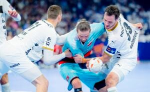 Drei Handballspieler kämpfen intensiv um den Ball; einer in einem blauen Trikot wird von zwei Gegnern in weißen Trikots während eines Spiels auf einem Hallenplatz festgehalten.
