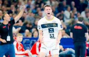 Ein männlicher Handballspieler in einem weiß-goldenen Trikot schreit leidenschaftlich auf dem Spielfeld, während eine andere Person, möglicherweise ein Trainer, im Hintergrund kräftig gestikuliert. Im unscharfen Hintergrund sind Zuschauer zu sehen.