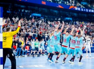 Eine Gruppe von Handballspielern in blauen Trikots blockt einen Schuss ab und hebt dabei die Arme, während ein gelb gekleideter Torwart zuschaut; im Hintergrund ist eine vollbesetzte Arena mit Zuschauern zu sehen.