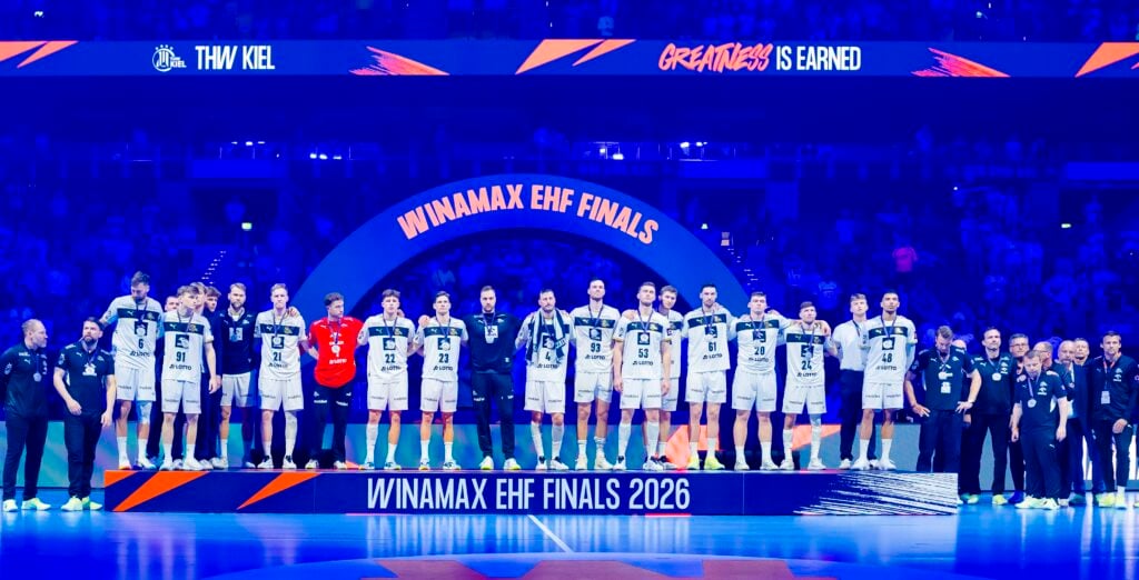 Eine Männerhandballmannschaft in weißen Trikots steht auf einem Podium mit der Aufschrift WINAMAX EHF FINALS 2026 in einer hell erleuchteten Arena, mit Trainern und Mitarbeitern auf jeder Seite und einem großen Bogen und einer Menschenmenge im Hintergrund.