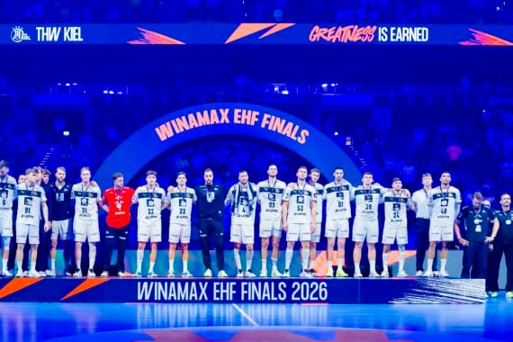 Eine Männerhandballmannschaft in weißen Trikots steht auf einem Podium mit der Aufschrift WINAMAX EHF FINALS 2026 in einer hell erleuchteten Arena, mit Trainern und Mitarbeitern auf jeder Seite und einem großen Bogen und einer Menschenmenge im Hintergrund.