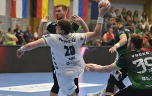 Ein Handballspieler in einem weißen Trikot springt, um den Ball zu schießen, während er während eines Spiels von Spielern in schwarzen und grünen Trikots verteidigt wird. Im Hintergrund sehen Zuschauer zu, und über dem Spielfeld hängen Fahnen.
