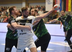 Ein Handballspieler in einem weißen Trikot versucht zu schießen, während zwei Gegner in grünen Trikots eng verteidigen. Die Szene findet in einer Halle statt, im Hintergrund sind Zuschauer und Fahnen zu sehen.