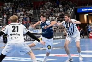 Drei männliche Handballspieler in Aktion während eines Spiels. Ein Spieler in einem dunkelblauen Trikot versucht zu passen oder zu schießen, während zwei gegnerische Spieler in weißen Trikots verteidigen. Eine Menschenmenge schaut von der Tribüne im Hintergrund zu.