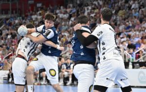 Vier männliche Handballspieler, zwei in blauen und zwei in weißen Trikots, sind auf dem Spielfeld in Aktion. Ein Spieler der blauen Mannschaft springt, während er den Ball hält, während im Hintergrund eine Menschenmenge zusieht.
