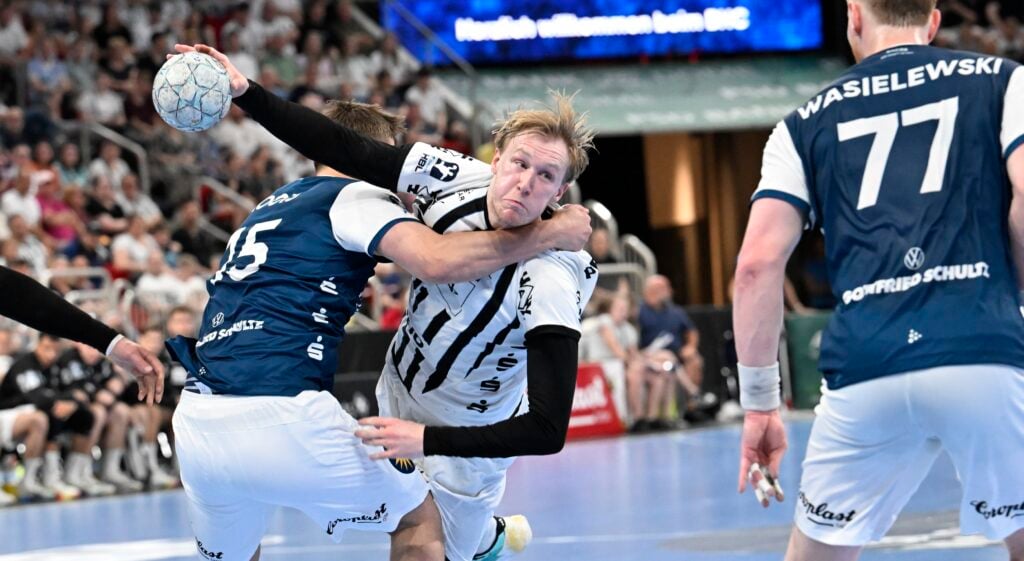 Ein Handballspieler in einem weiß-schwarzen Trikot versucht zu schießen, während er von zwei Spielern in blauen Trikots während eines rasanten Spiels verteidigt wird, während die Zuschauer im Hintergrund zuschauen.