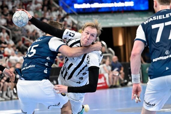 Ein Handballspieler in einem weiß-schwarzen Trikot versucht zu schießen, während er von zwei Spielern in blauen Trikots während eines rasanten Spiels verteidigt wird, während die Zuschauer im Hintergrund zuschauen.