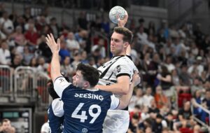 Ein Handballspieler in einem weiß-schwarzen Trikot springt zum Schuss, während zwei Verteidiger in blauen Trikots, eines davon mit der Nummer 49, versuchen, ihn abzuwehren. Die Zuschauer auf der Tribüne verfolgen das intensive Spiel.