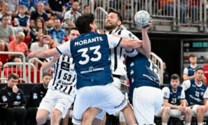 Zwei Handballspieler in blau-weißen Trikots verteidigen sich gegen einen gegnerischen Spieler in einem weiß-schwarzen Trikot, der den Ball hält, während die Zuschauer im Hintergrund das intensive Geschehen beobachten.
