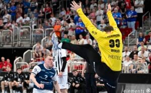 Ein Handballtorwart in einem gelben Trikot springt vor, um einen Schuss abzublocken, und breitet Arme und Beine aus, während ein Spieler in einem blau-weißen Trikot den Ball ins Tor schießt. Die Zuschauer füllen die Tribünen im Hintergrund.