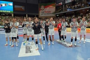 Eine Männer-Handballmannschaft in schwarz-weiß gestreiften Trikots steht gemeinsam auf einem Spielfeld, einige klatschen, während im Hintergrund einer Halle ein großes Publikum und Spruchbänder zu sehen sind.