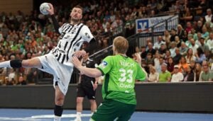 Ein Handballspieler in einem weiß-schwarzen Trikot springt, um den Ball zu werfen, während ein Verteidiger in einem grünen Trikot mit der Reichszahl 33 auf dem Rücken ihn blockiert. Die Aktion findet vor einer Menschenmenge in einer Halle statt.