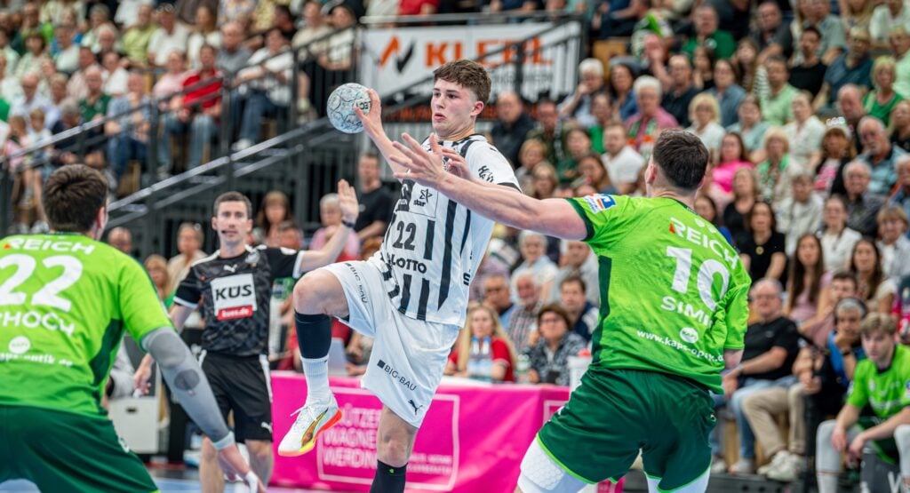 Ein Handballspieler in einem weiß-schwarzen Trikot springt, um den Ball zu werfen, während zwei Verteidiger in Grün versuchen, ihn zu blockieren. Das Spiel findet in einer Halle statt und wird von der Tribüne aus verfolgt.