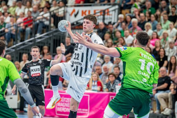 Ein Handballspieler in einem weiß-schwarzen Trikot springt, um den Ball zu werfen, während zwei Verteidiger in Grün versuchen, ihn zu blockieren. Das Spiel findet in einer Halle statt und wird von der Tribüne aus verfolgt.