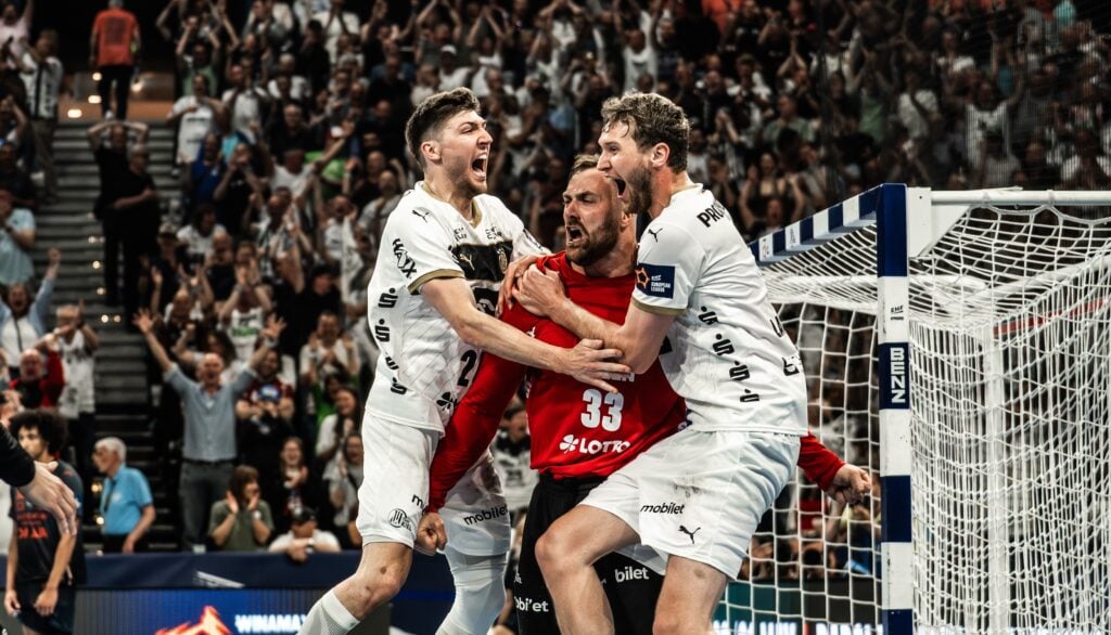 26_nach_MHB_TitelJubel Drei männliche Handballspieler in weißen Trikots feiern leidenschaftlich vor einem Torpfosten, mit einer jubelnden Menge im Hintergrund. Der Spieler in der Mitte trägt ein rotes Trikot und schreit vor Freude, während seine Mannschaftskameraden ihn umarmen.