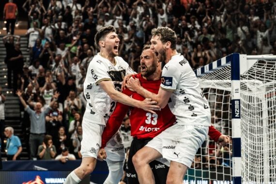 Drei männliche Handballspieler in weißen Trikots feiern leidenschaftlich vor einem Torpfosten, mit einer jubelnden Menge im Hintergrund. Der Spieler in der Mitte trägt ein rotes Trikot und schreit vor Freude, während seine Mannschaftskameraden ihn umarmen.