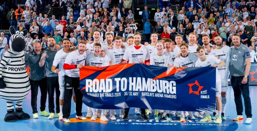 Ein jubelndes Handballteam posiert auf einem Spielfeld, lächelnd und mit einem Road to Hamburg EHF Finals-Banner in der Hand, mit einem Zebra-Maskottchen und jubelnden Fans im Hintergrund.