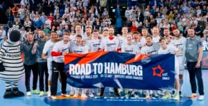 Ein jubelndes Handballteam posiert auf einem Spielfeld, lächelnd und mit einem Road to Hamburg EHF Finals-Banner in der Hand, mit einem Zebra-Maskottchen und jubelnden Fans im Hintergrund.